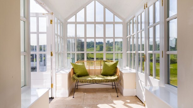 A conservatory with a high ceiling, large windows, a wicker chair with cushions, and a view of a green landscape. - Grass Ings (Ambleside)