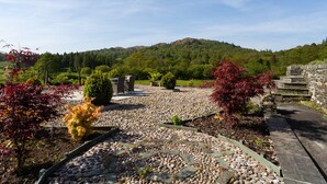 A garden with a stone pathway, surrounded by trees and a mountain in the background. - Grass Ings (Ambleside)