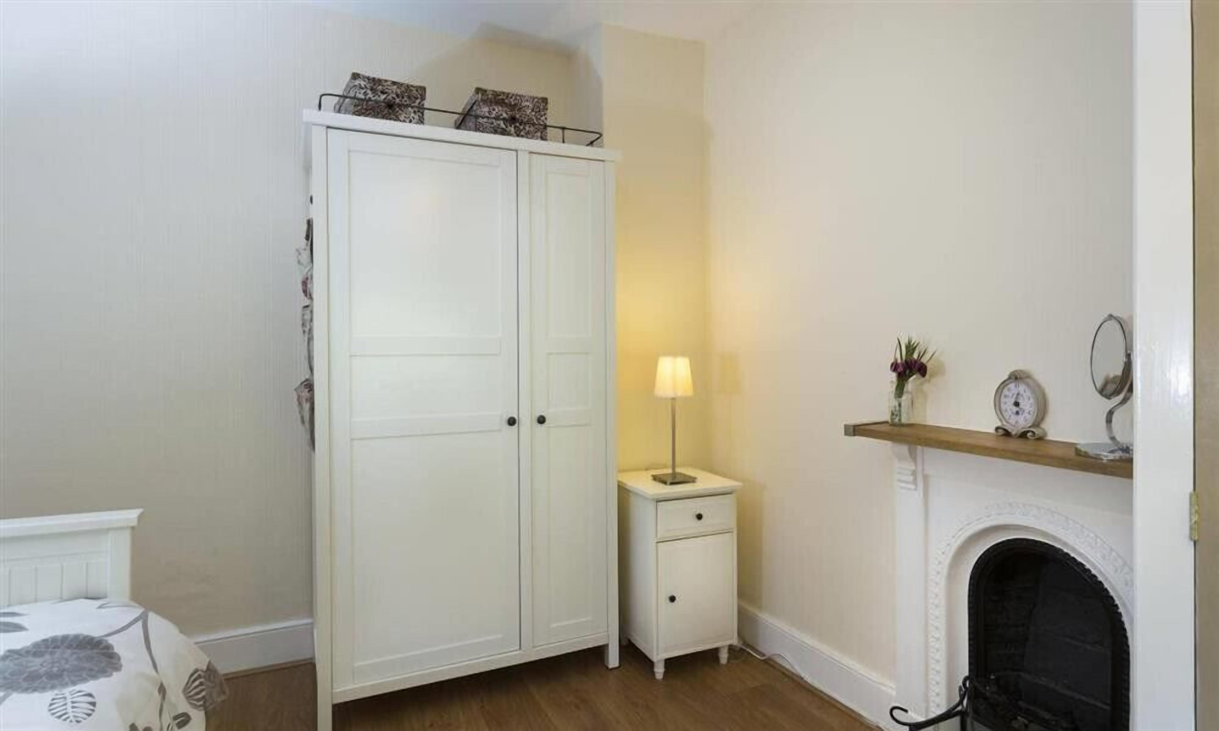 Une pièce avec une armoire blanche, une table de chevet avec une lampe et une cheminée.