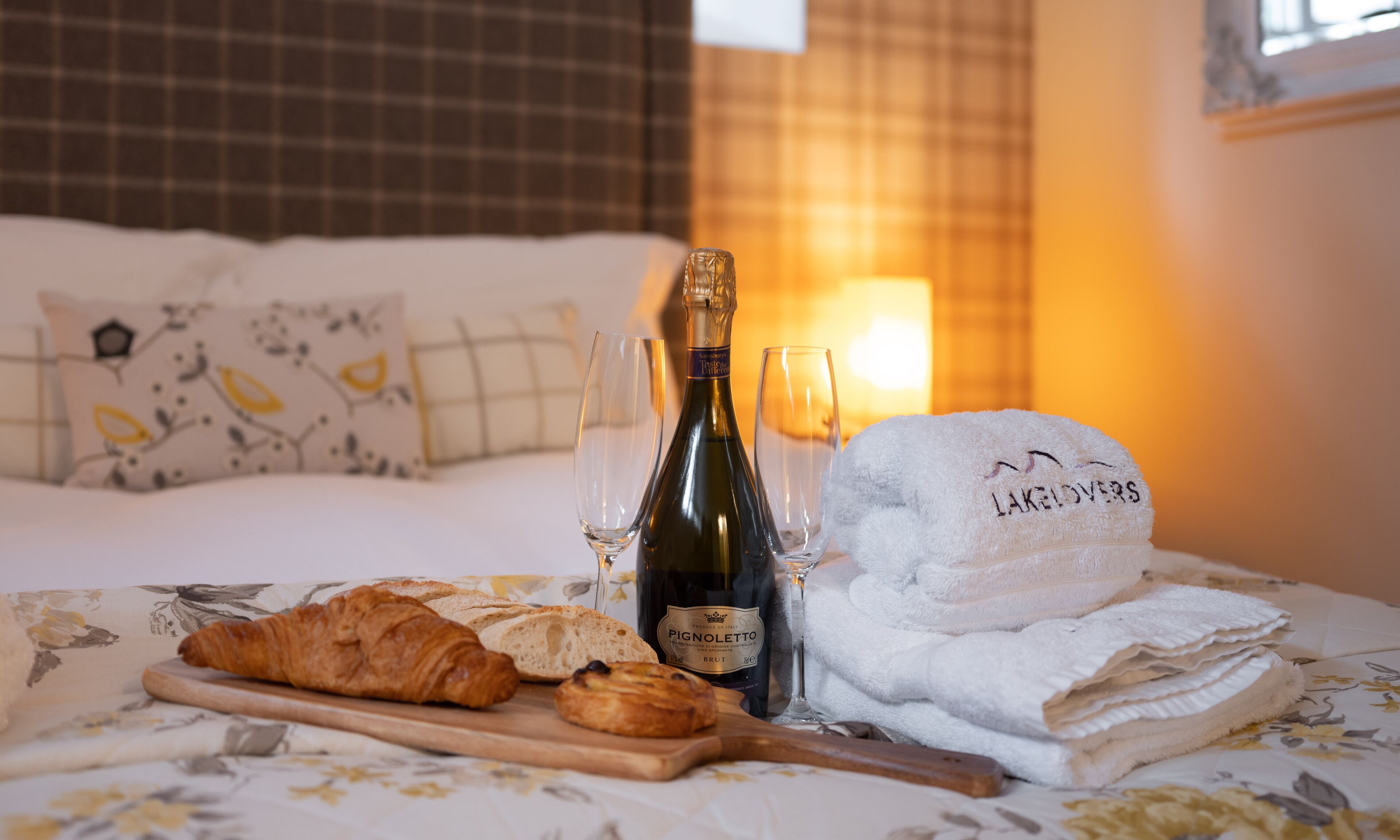 A bed with a floral bedspread, a bottle of Pignoletto wine, two glasses, a croissant, and a towel with "LAKELOVERS" on it.
