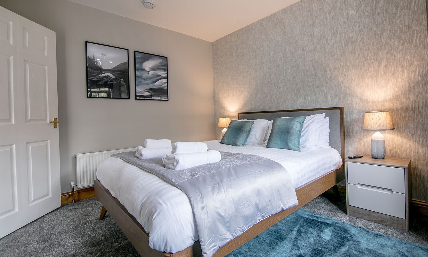 Un lit bien fait, avec du linge de lit blanc et un tapis bleu qui apporte une touche de couleur, une lampe de chevet et deux tableaux encadrés accrochés au mur.