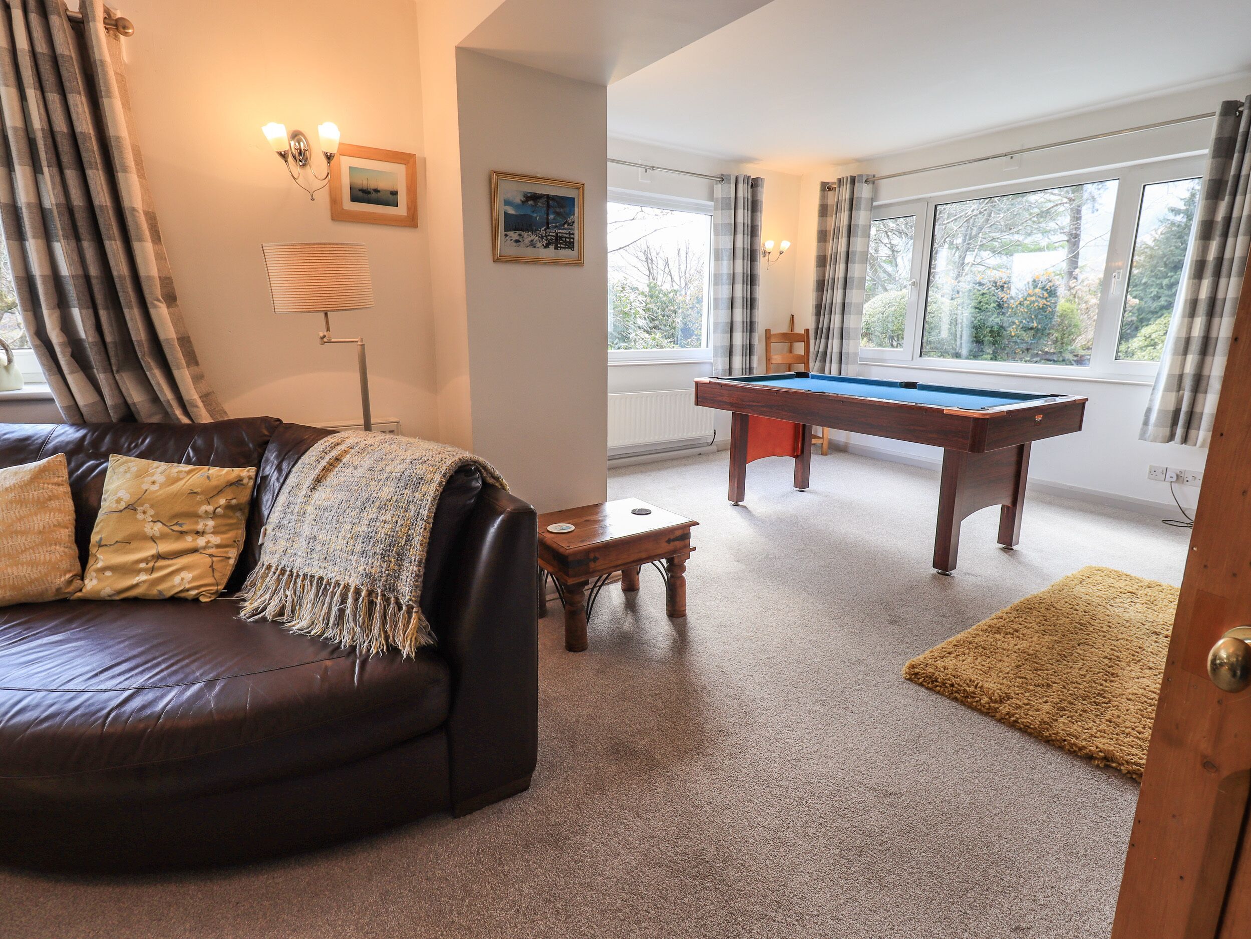 A cozy living room with a dark leather sofa, a pool table, and a view through large windows.
