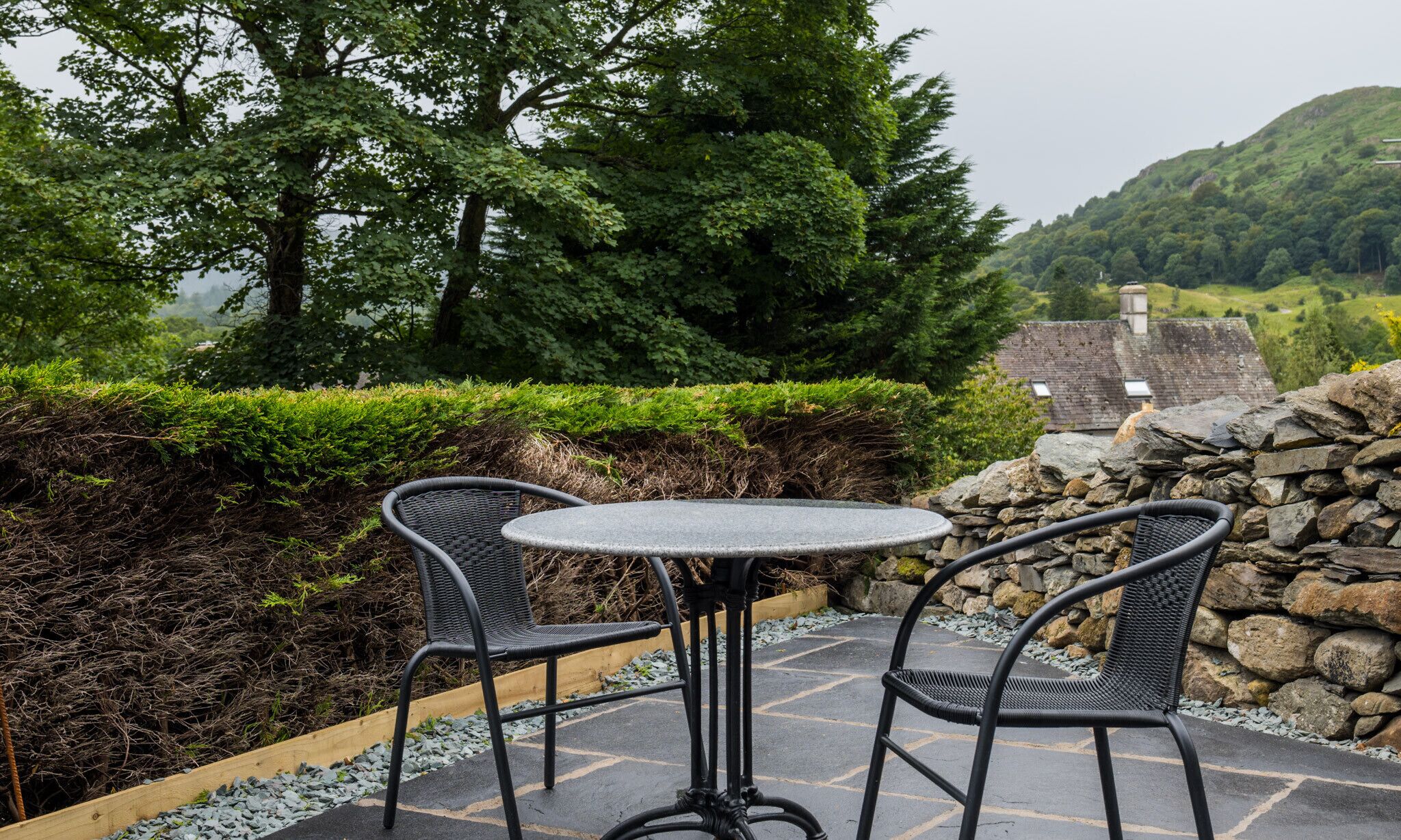 Un patio con dos sillas de mimbre y una mesa redonda, rodeado por un muro de piedra y un seto, con vistas a un paisaje rural con árboles y una casa.