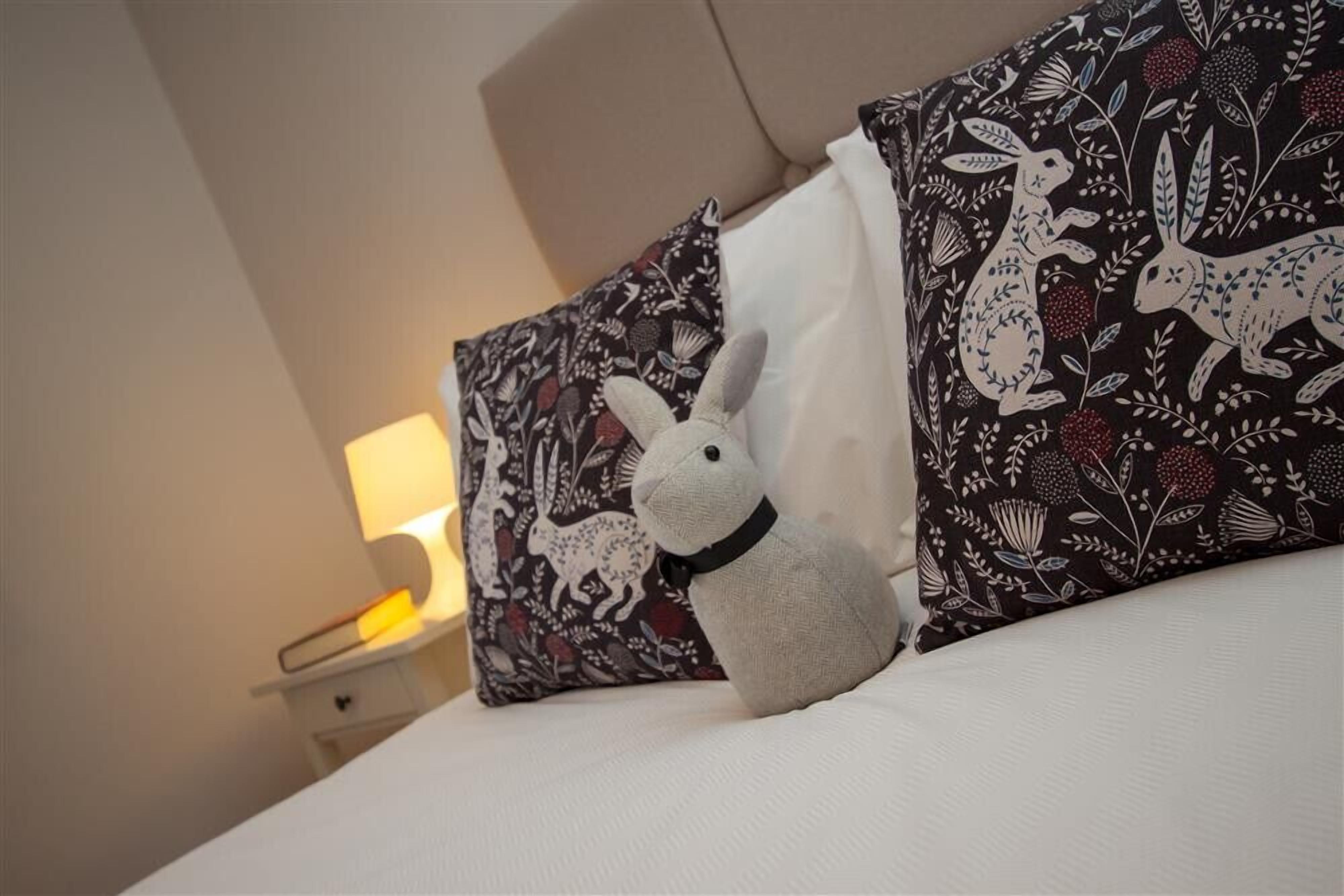 Una cama con dos almohadones decorativos, uno con estampado floral y otro con diseño de conejo blanco, y un pequeño peluche de conejo sobre la cama.
