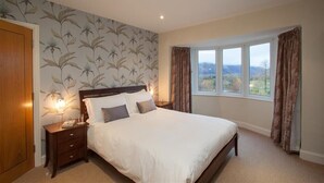 A bedroom with a large bed, a nightstand, a lamp, and a window with curtains. - Riggside (Keswick)