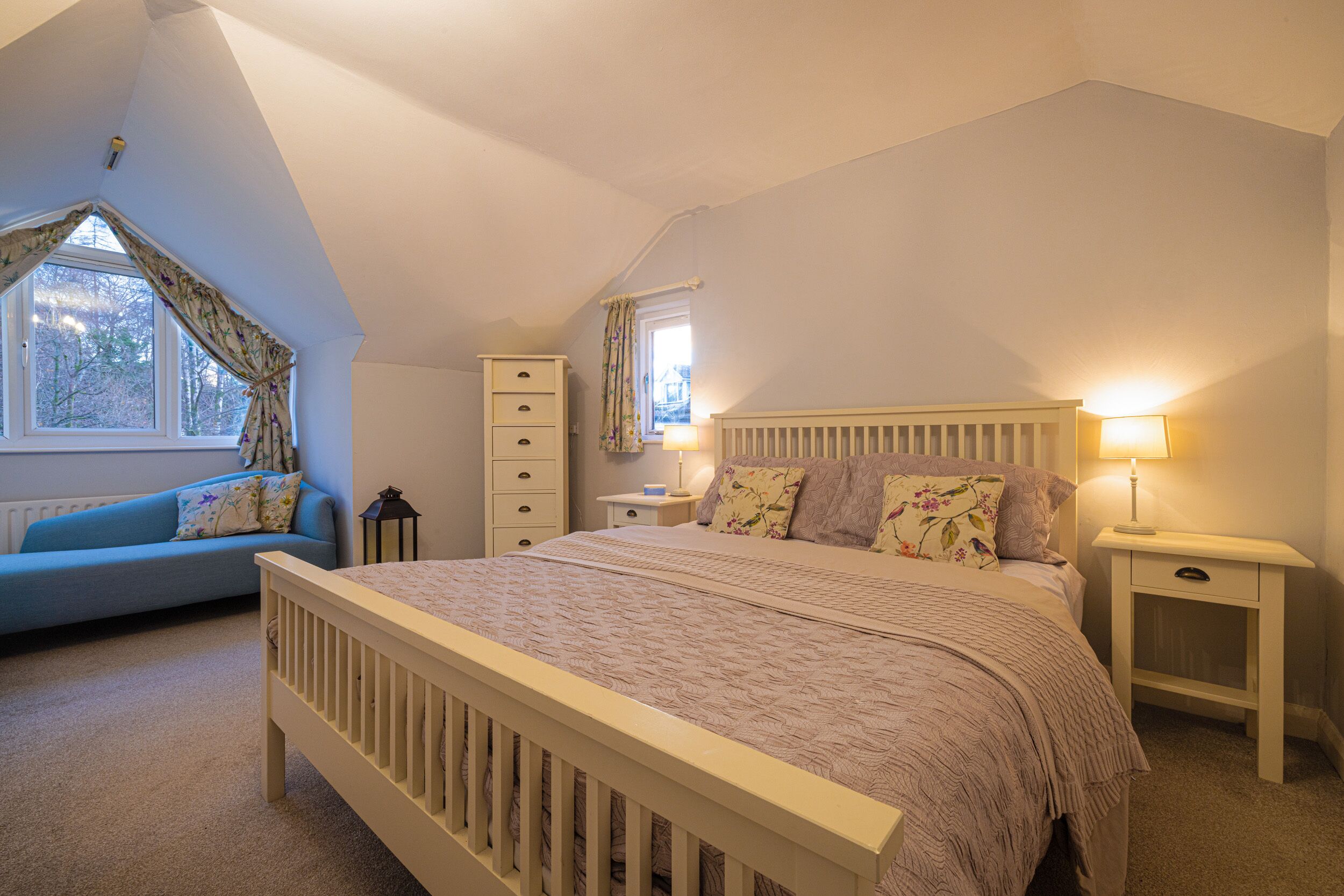 A bedroom with a large bed, a dresser, a nightstand, a lamp, a window with curtains, and a blue sofa.