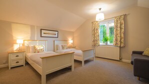 A bedroom with two single beds, bedside tables with lamps, a radiator, a window with floral curtains, and a grey sofa. - Holly Cottage (Ambleside)