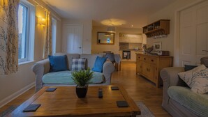 A cozy living room with a wooden coffee table, two sofas with blue cushions, a potted plant, and a kitchen area in the background. - Holly Cottage (Ambleside)