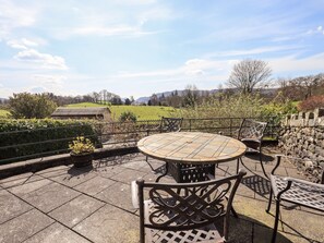 An outdoor patio with a round table and chairs, stone walls, and a view of a green field and hills. - Oak Bank (Windermere)