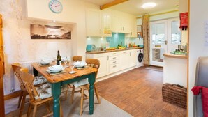 Un coin repas avec une table en bois dressée pour quatre personnes, une horloge au mur et une cuisine dotée de meubles blancs et d’un lave-linge.