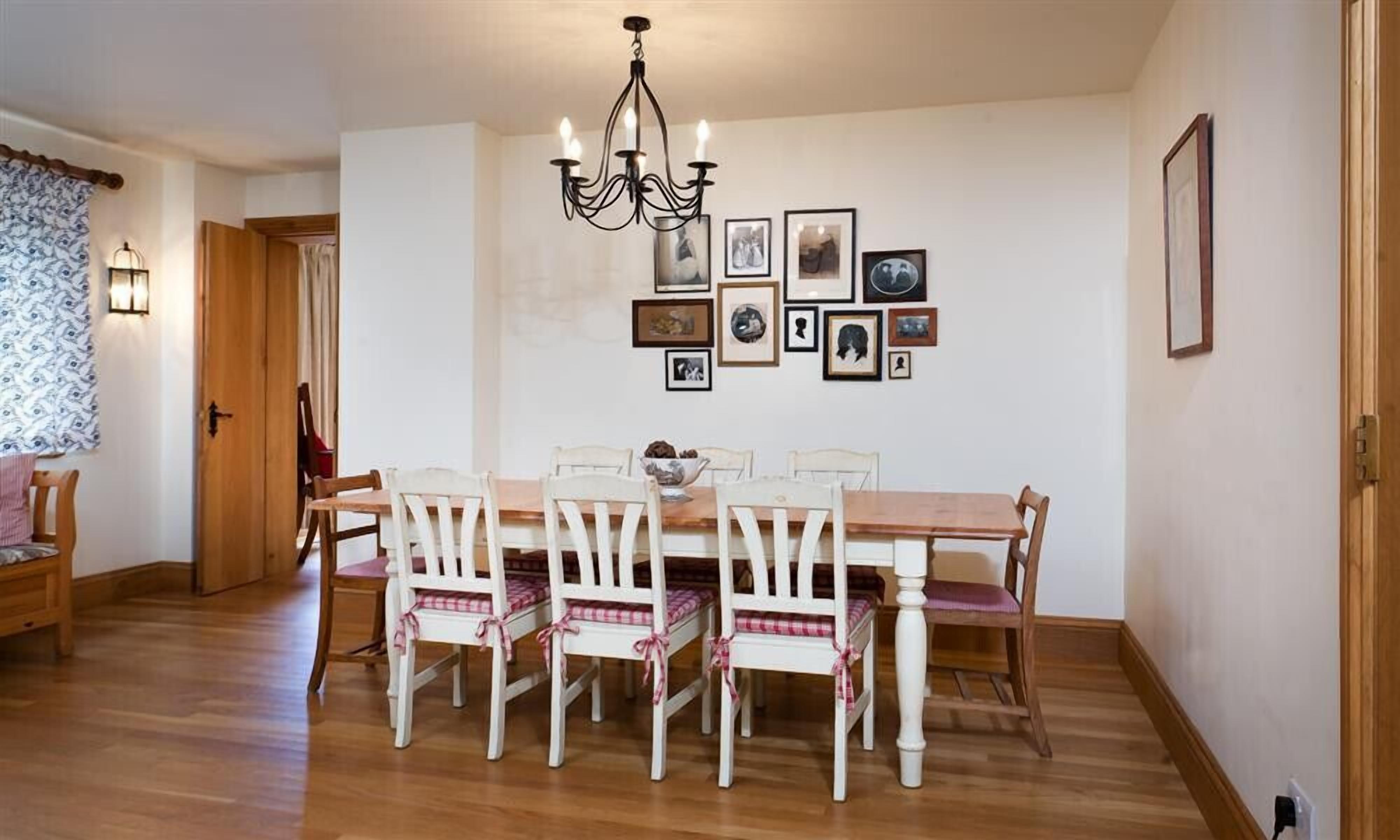 Un comedor con mesa de madera y sillas blancas, una araña y cuadros enmarcados en la pared.