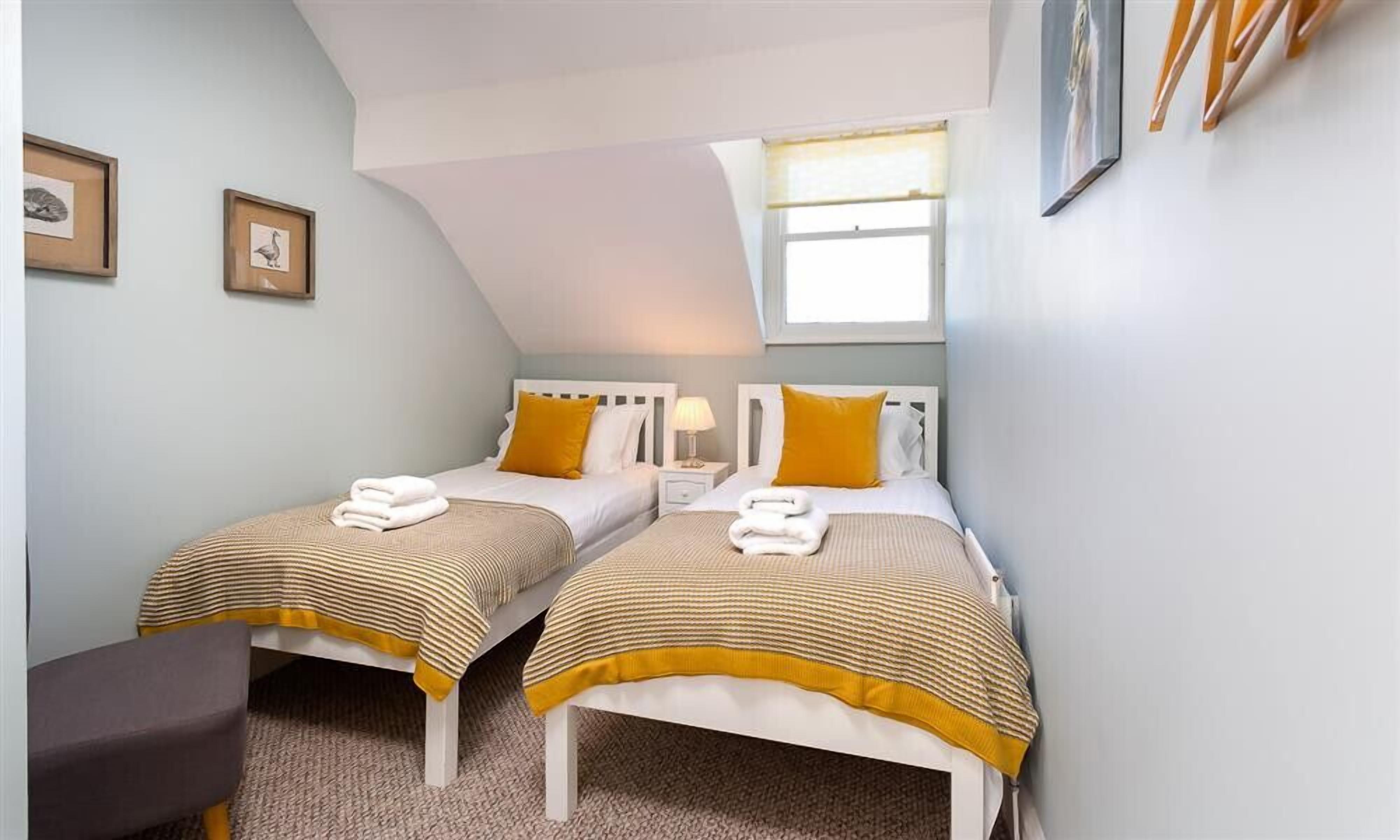 A bedroom with two beds, each with a striped bedspread and a yellow pillow. There is a nightstand with a lamp between the beds, and a chair in the corner.