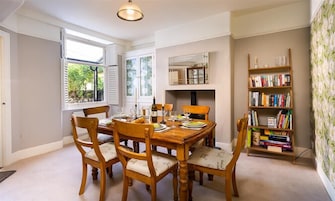 Uma sala de jantar com uma mesa de madeira posta para seis pessoas, rodeada por seis cadeiras. Há uma estante à direita com vários livros e objetos decorativos.