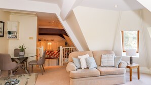 A modern living room with a glass coffee table, a beige sofa with cushions, a dining area with a glass table, and a kitchen in the background. - Loughrigg Suite (Ambleside)