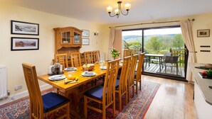 A dining area with a wooden table set for six, surrounded by chairs with blue cushions. A wooden cabinet with glass doors is against the wall, and there are framed pictures on the wall.