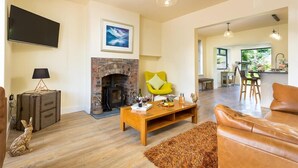 A living room with a fireplace, a television, a bright yellow chair, a leather sofa, and a kitchen area in the background. - Greengarth (Keswick)