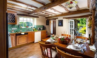 A rustic kitchen with wooden beams, a dining area with a table set for six, and a view of greenery outside.
