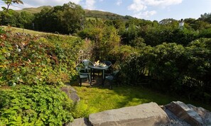 Un jardín con una mesa y sillas, rodeado de abundante vegetación y colinas al fondo.