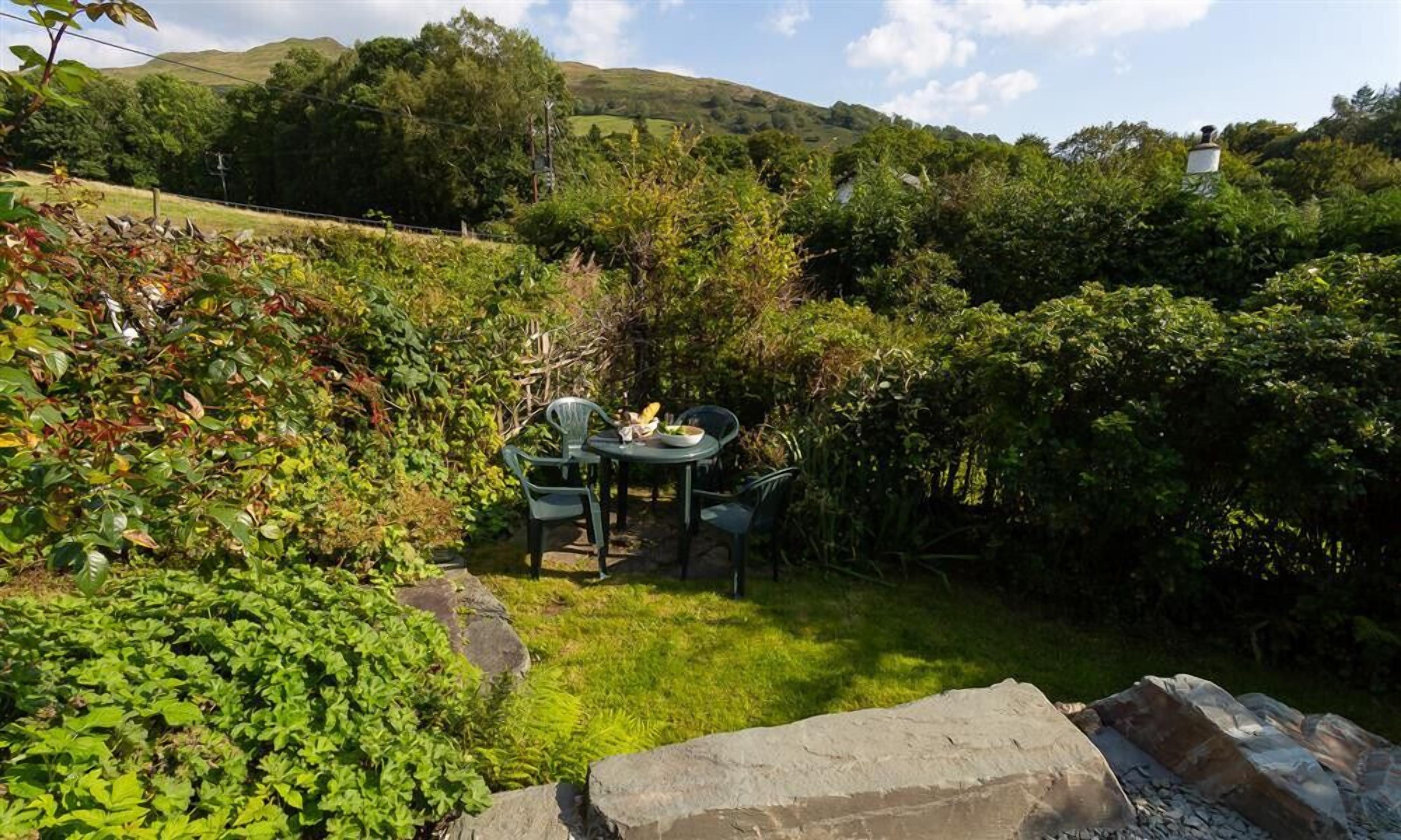 A garden with a table and chairs, surrounded by lush greenery and hills in the background.