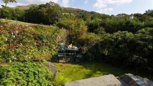 Un jardín con una mesa y sillas, rodeado de abundante vegetación y colinas al fondo.