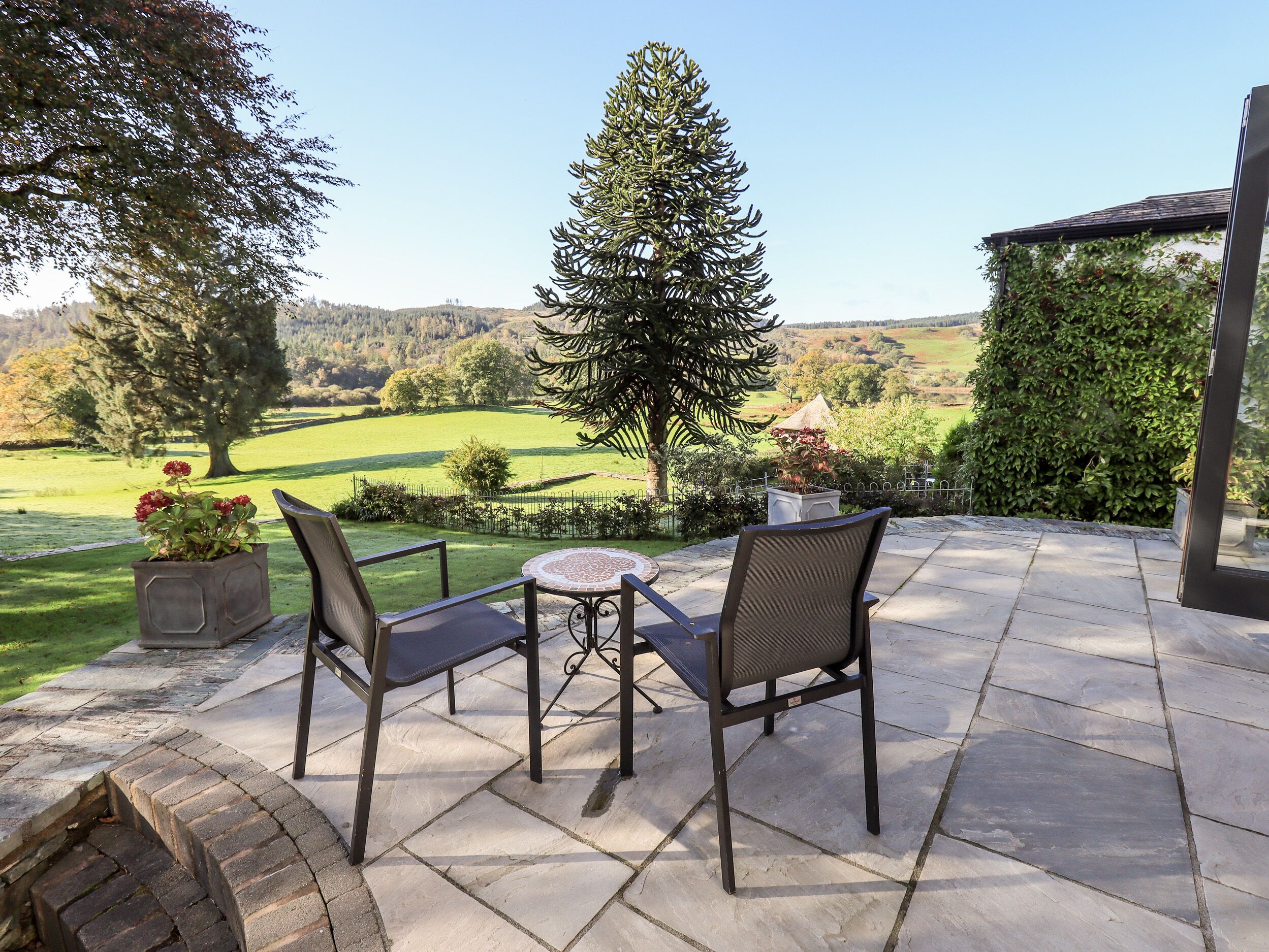 An outdoor patio with a table and chairs, surrounded by a stone floor and a view of a green landscape.