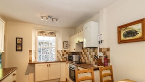 A compact kitchen with white cabinets, a microwave, a stove, and a window with a view of a stone wall. There are wooden chairs and a table with a bowl. - Jessicas Cottage (Windermere)
