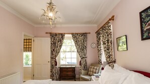 A bedroom with a bed, a nightstand, a chair, a window with curtains, a chandelier, and a clock on the wall. - Jessicas Cottage (Windermere)