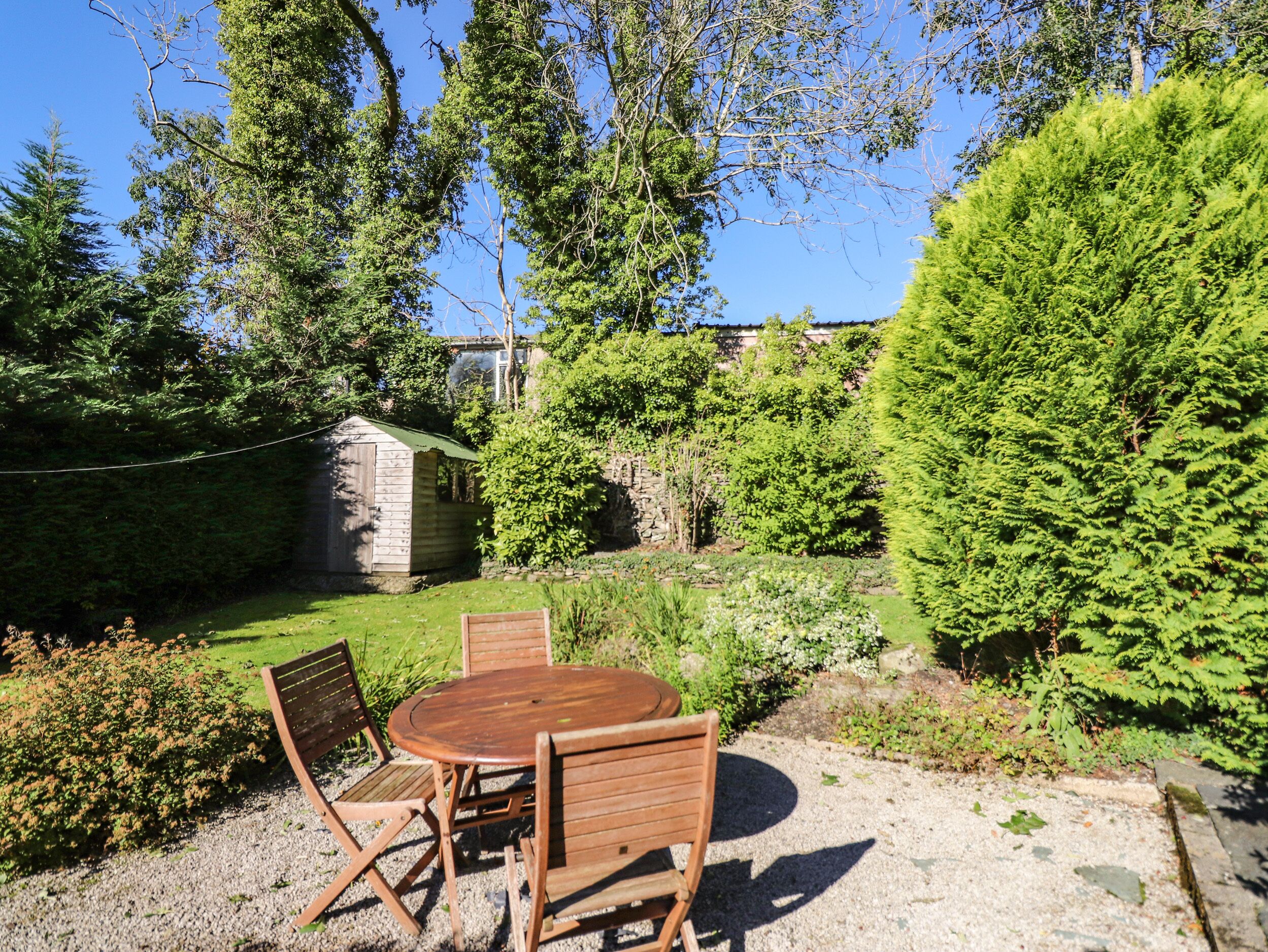 A wooden table and chairs set on a gravel surface in a garden with a shed and dense greenery.
