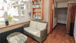 Une pièce chaleureuse avec un canapé en osier, une petite table en verre, une bibliothèque en bois et une vue dégagée sur l’extérieur grâce à une grande fenêtre.