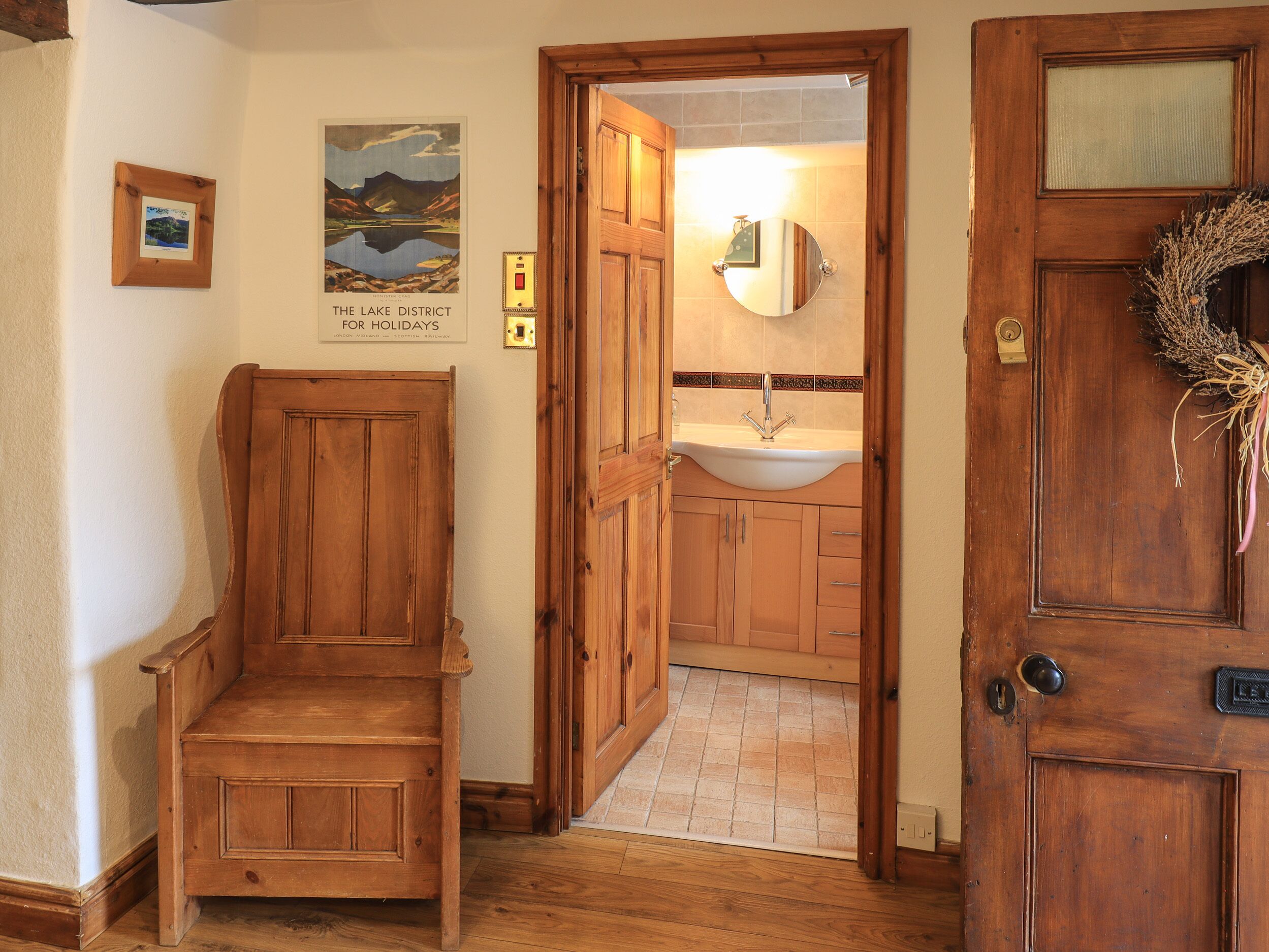 Une chaise en bois dans un couloir, une porte en bois ornée d’une guirlande, et une salle de bain avec un lavabo et un miroir.