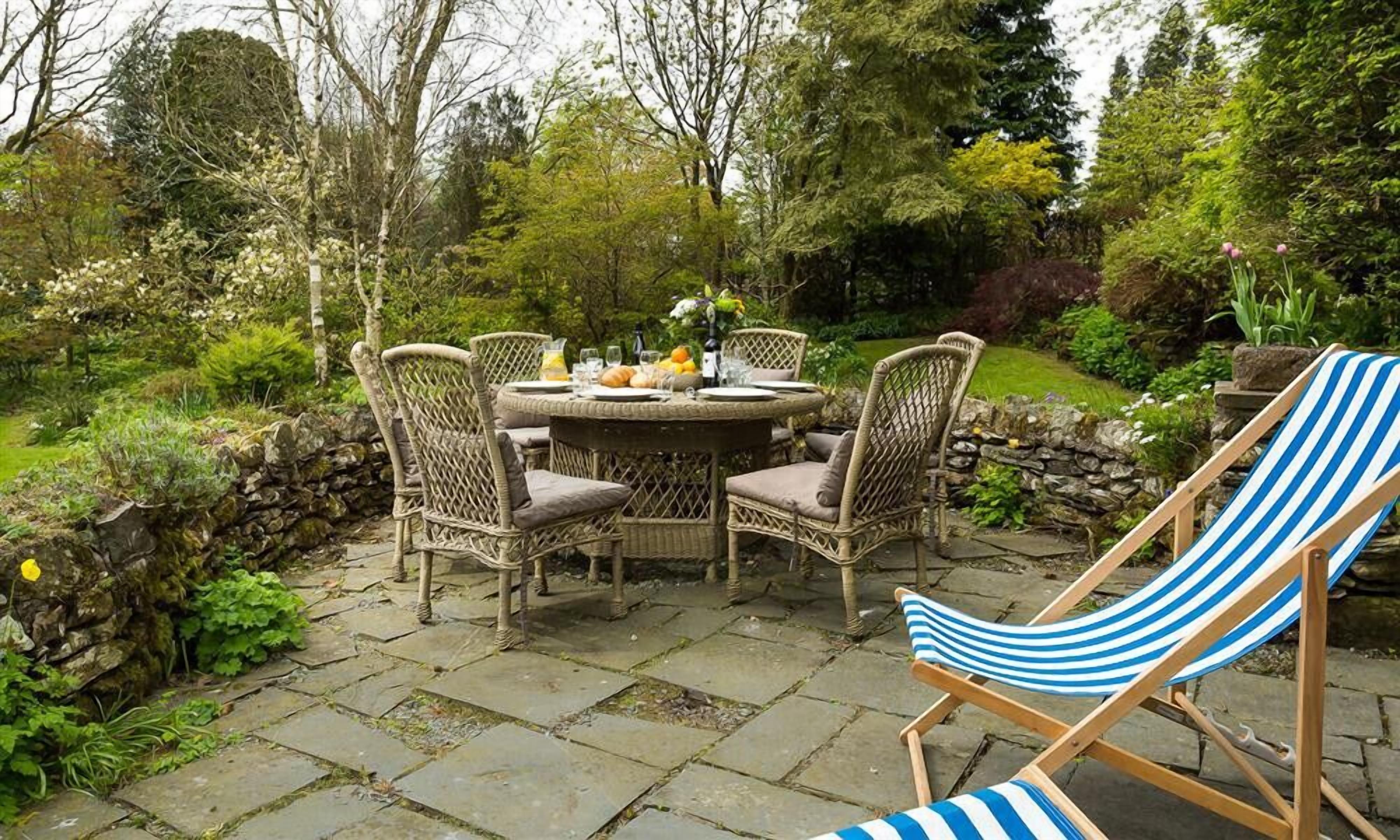 A round outdoor dining table with wicker chairs, surrounded by a stone wall and lush greenery.