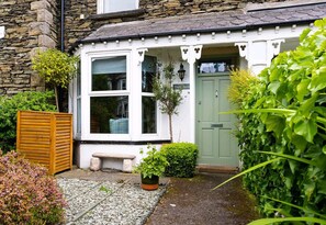 A quaint cottage with a stone facade, a green door, and a small garden area with a bench and potted plants. - Fern Bank Cottage (Windermere)