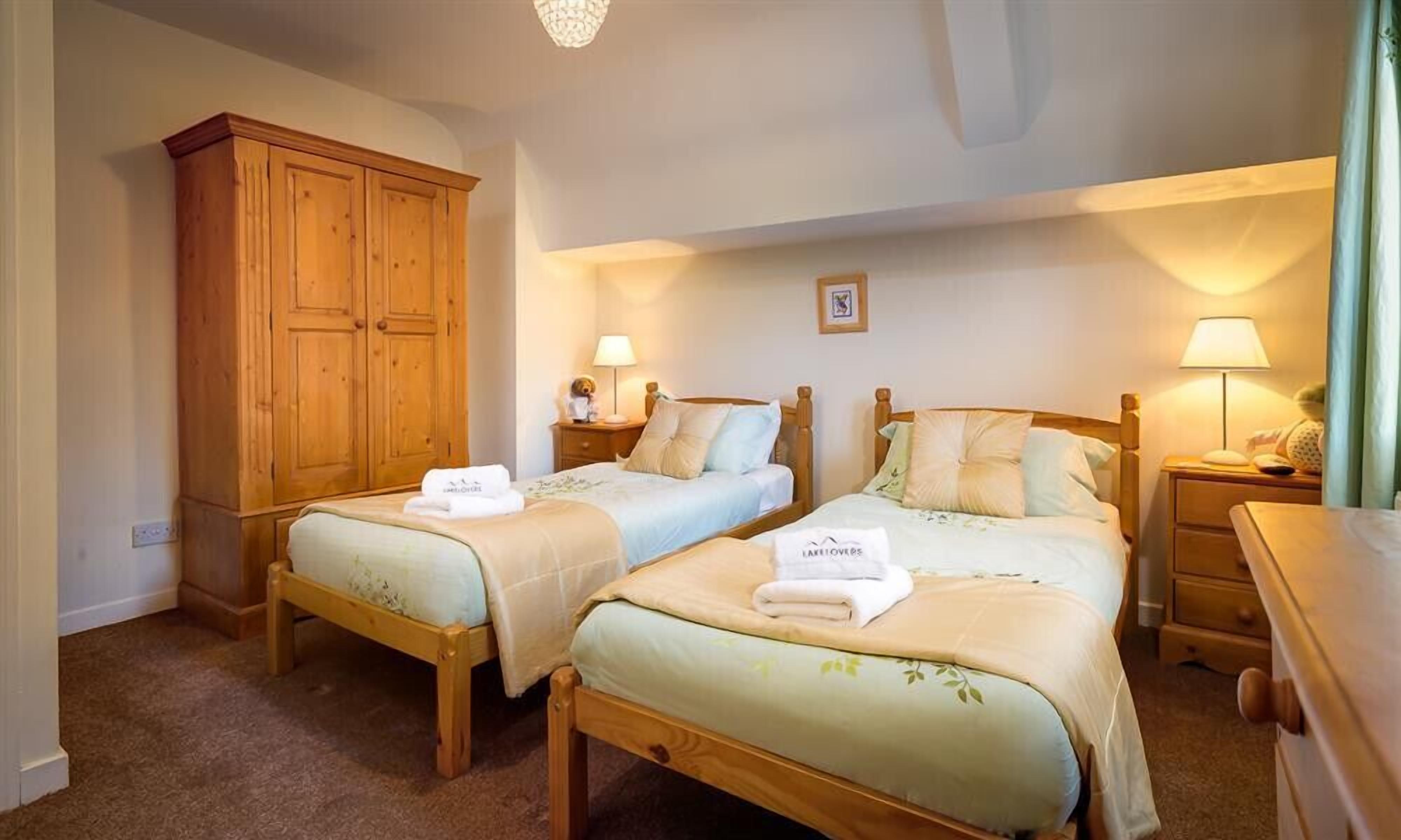 A hotel room with two wooden beds, each with a bedside table and lamp. There is a wooden wardrobe and a small framed picture on the wall.