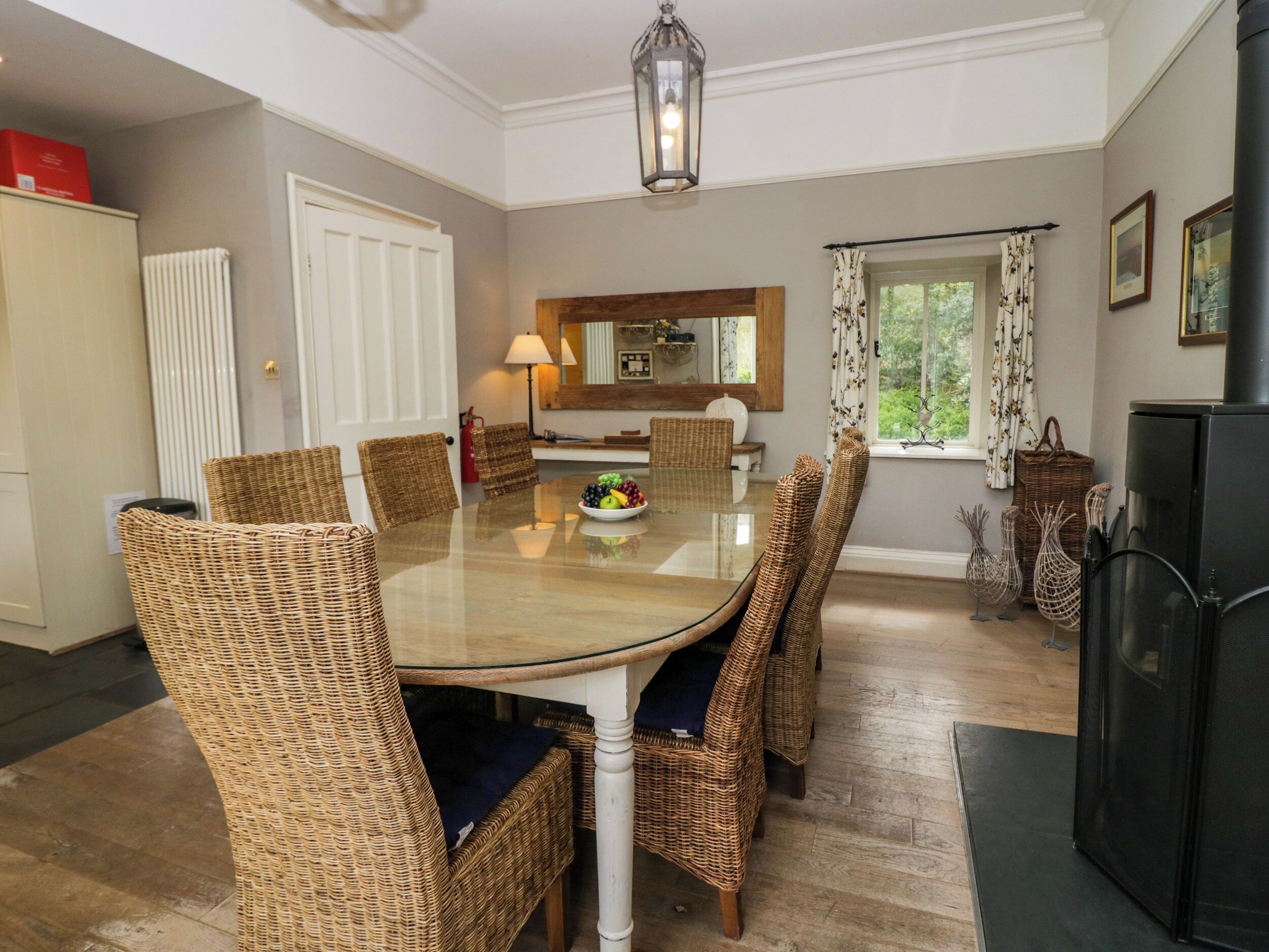 A dining area with a glass table surrounded by wicker chairs, a kitchen in the background, and a window with curtains.