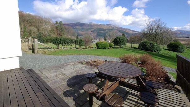 Una terrazza in legno con tavolo da picnic e panchine, circondata da un vialetto sterrato e vista su dolci colline e alberi.