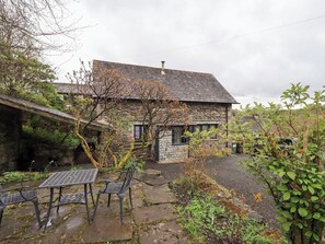 Une maison en pierre avec un toit en pente, un petit jardin avec une table et des chaises, et une voiture garée à proximité.