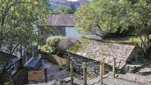 Un sendero de piedra que lleva a una terraza de madera con vistas a un lago, rodeado de vegetación exuberante y muros de piedra.