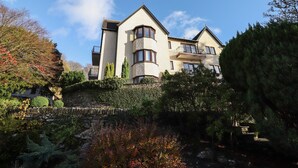 A multi-story building with a stone retaining wall, multiple balconies, and a garden area with various plants. - Romney 14 (Ambleside)