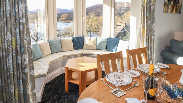 A dining area with a wooden table set for two, a view of a lake and hills through large windows, and a cozy seating area with a curved bench. - Romney 14 (Ambleside)