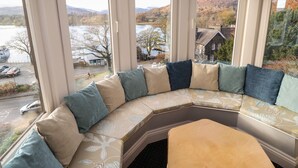 A curved bench with multiple cushions, large windows, and a view of a lake and buildings. - Romney 14 (Ambleside)