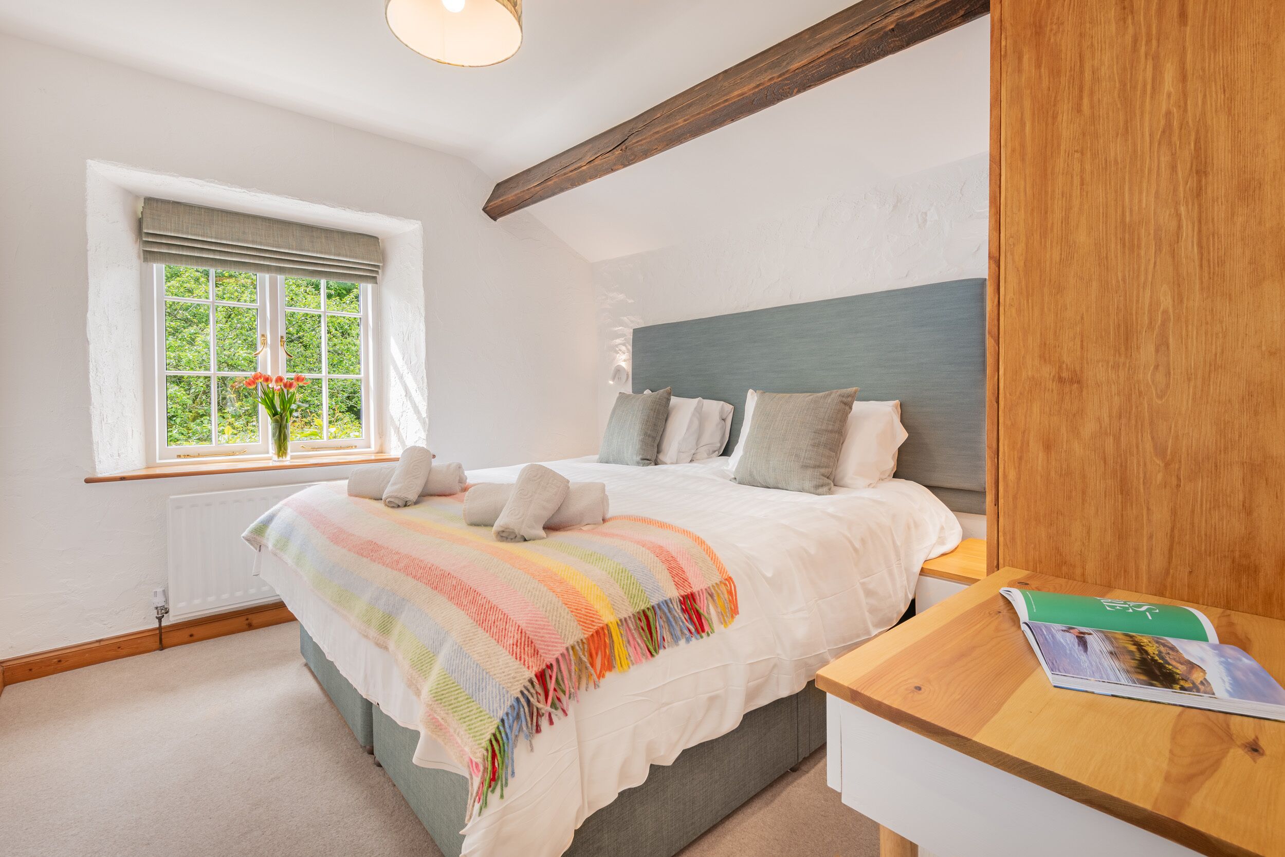 Un dormitorio con una cama grande, una ventana con vista a follaje, cabecera de madera y una mesita de noche con un libro.