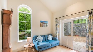 Une pièce avec un canapé bleu, un meuble en bois, une petite table avec une lampe, une grande fenêtre et une porte donnant sur un espace ext érieur agrémenté d’un mur en pierre et de plantes.
