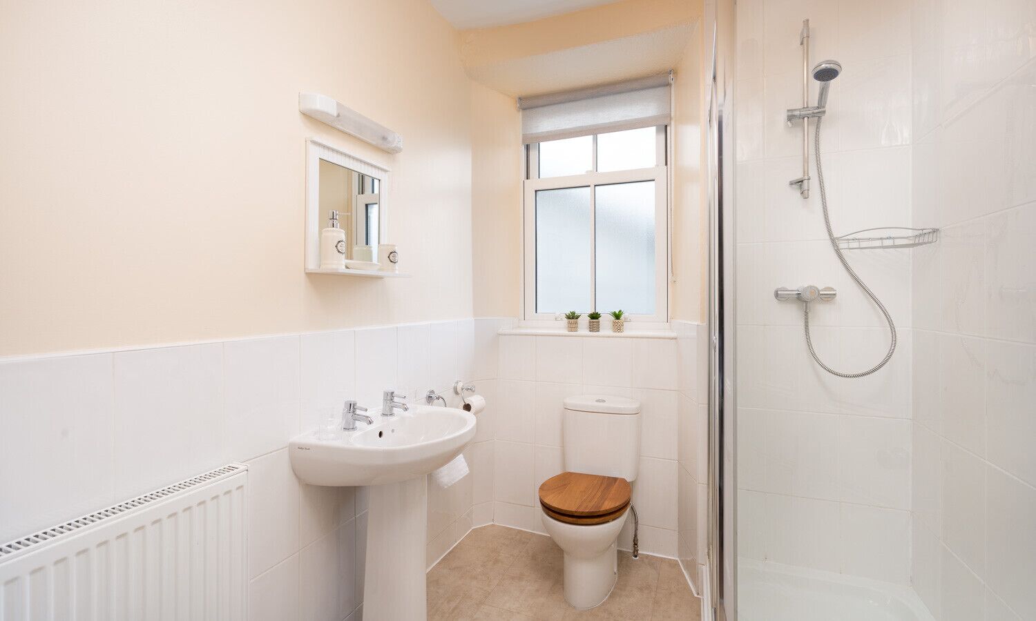 A small bathroom with a white sink, a wooden toilet seat, a window, and a shower.