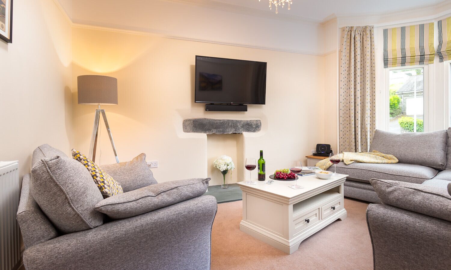 A modern living room with a flat-screen TV mounted on the wall, a white coffee table with a wine bottle and glasses, and two grey sofas.