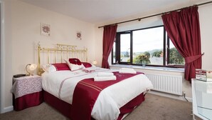 A hotel room with a bed, a bedside table, a lamp, a window with curtains, and a view of the outdoors. - Fairhaven (Windermere)