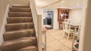 Eine Treppe führt zu einem Essbereich mit Tisch und Stühlen, im Hintergrund ist eine Küche zu sehen und ein Holzregal mit Geschirr.