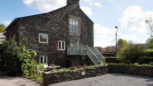 A stone building with a staircase leading to a balcony, surrounded by greenery and a gravel driveway. - Glen View Cottage (Ambleside)
