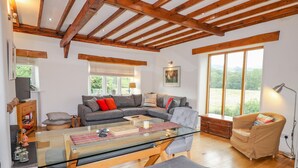 A spacious living room with a wooden ceiling, a grey sectional sofa, a glass coffee table, and a large window offering a view of greenery. - Glen View Cottage (Ambleside)