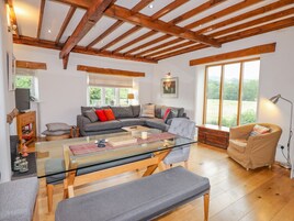 Amplia sala de estar con techo de madera, un sofá seccional gris, una mesa de centro de vidrio y un ventanal con vistas a follaje.