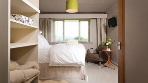 A hotel room with a bed, a chair, a small table with a vase, a television, and a window with curtains. - Harrison Stickle (Windermere)